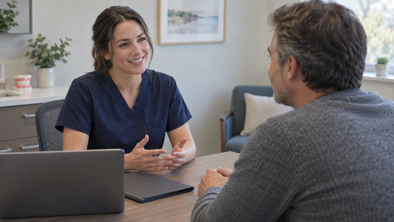 Dental team member discusses treatment with a patient across a desk with a laptop and closed folder.